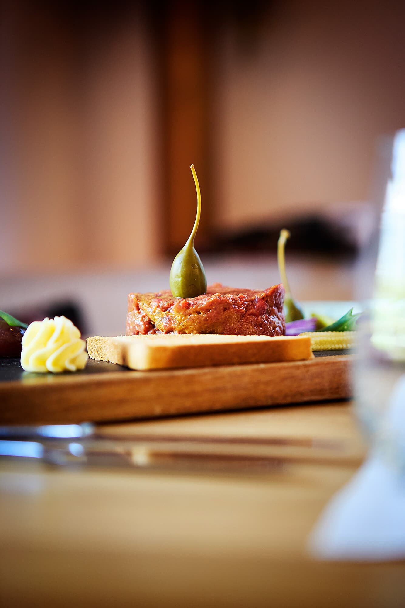 An elegant dish on a wooden board with beef tartare garnished with a caper berry and a small portion of butter at the B Smart Hotel Widnau.