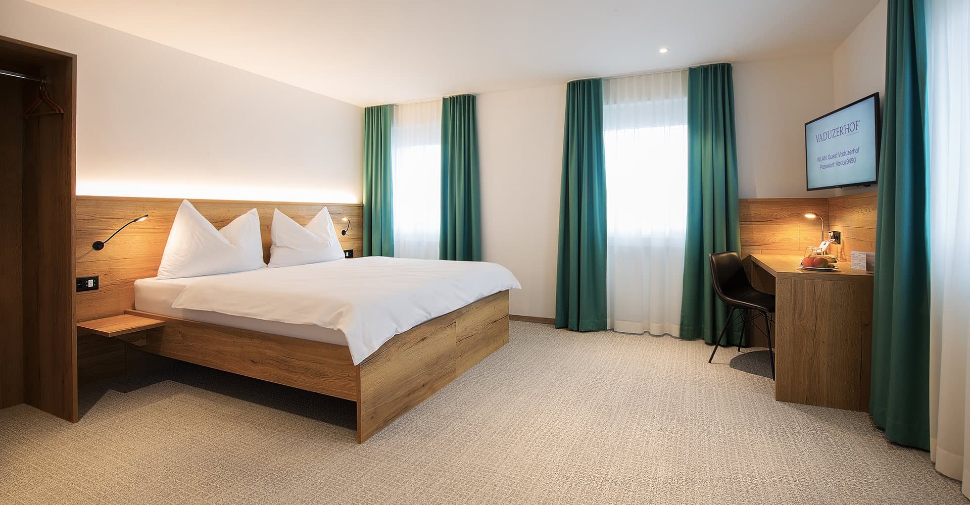 A modern hotel room in the Vaudzerhof with a large bed, wooden furniture, blue-green curtains, and a desk with a chair.