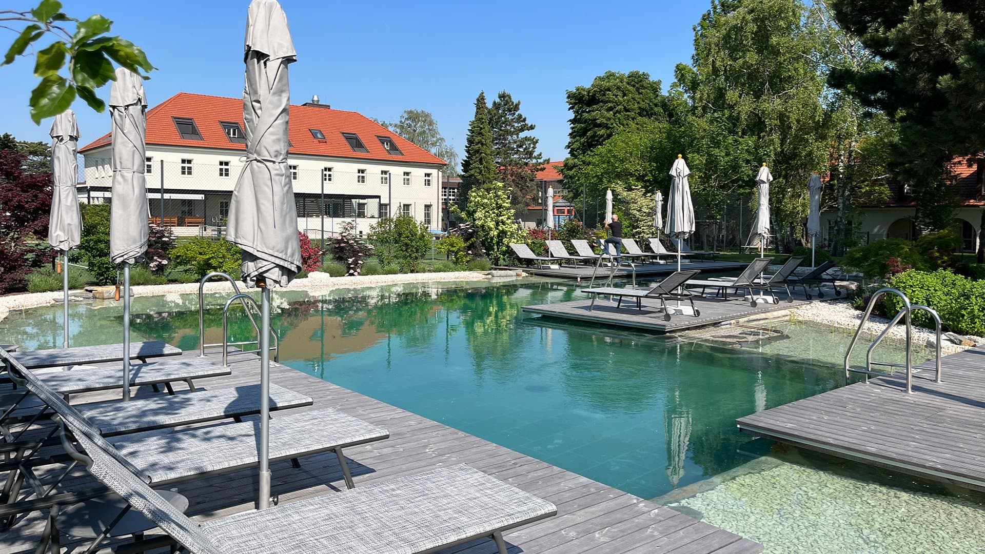 Outdoor pool of the Hotel Spitzerberg by b_smart with lounge chairs and closed umbrellas, surrounded by green landscape and a building with a red roof in the background.