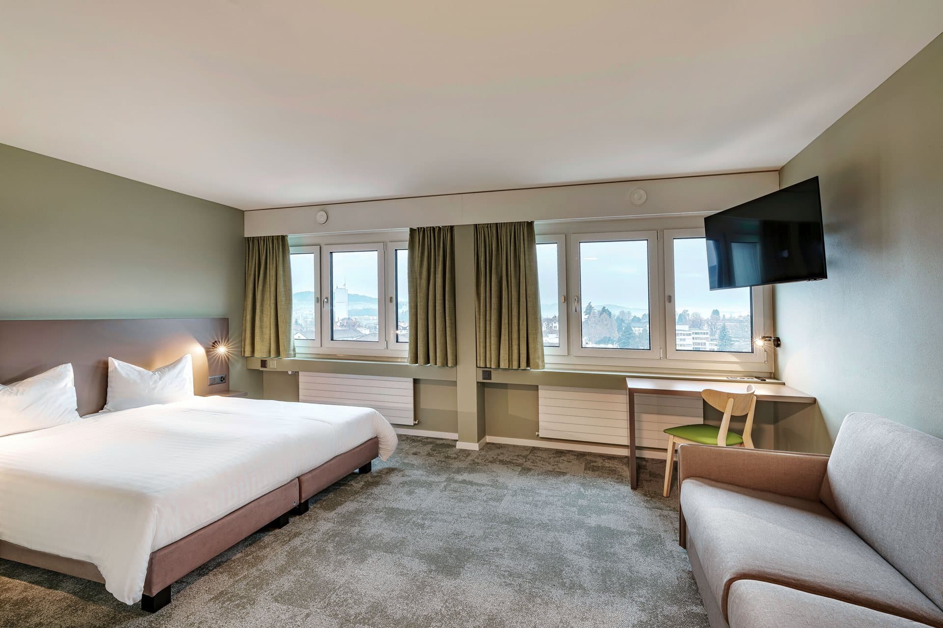 A modern hotel room with a large bed, a sofa, a desk with a chair, a wall-mounted TV, and several windows with green curtains in the Flexy Motel Wil Junior Suite.