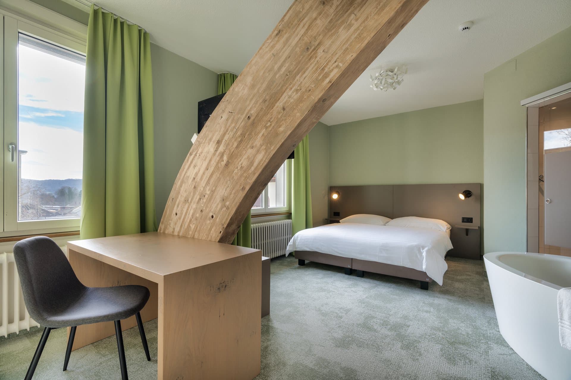 The room in the Flexy Motel Schonenwerd is modernly furnished with a double bed, a wooden workspace, and a freestanding bathtub, with green walls and curtains defining the space.