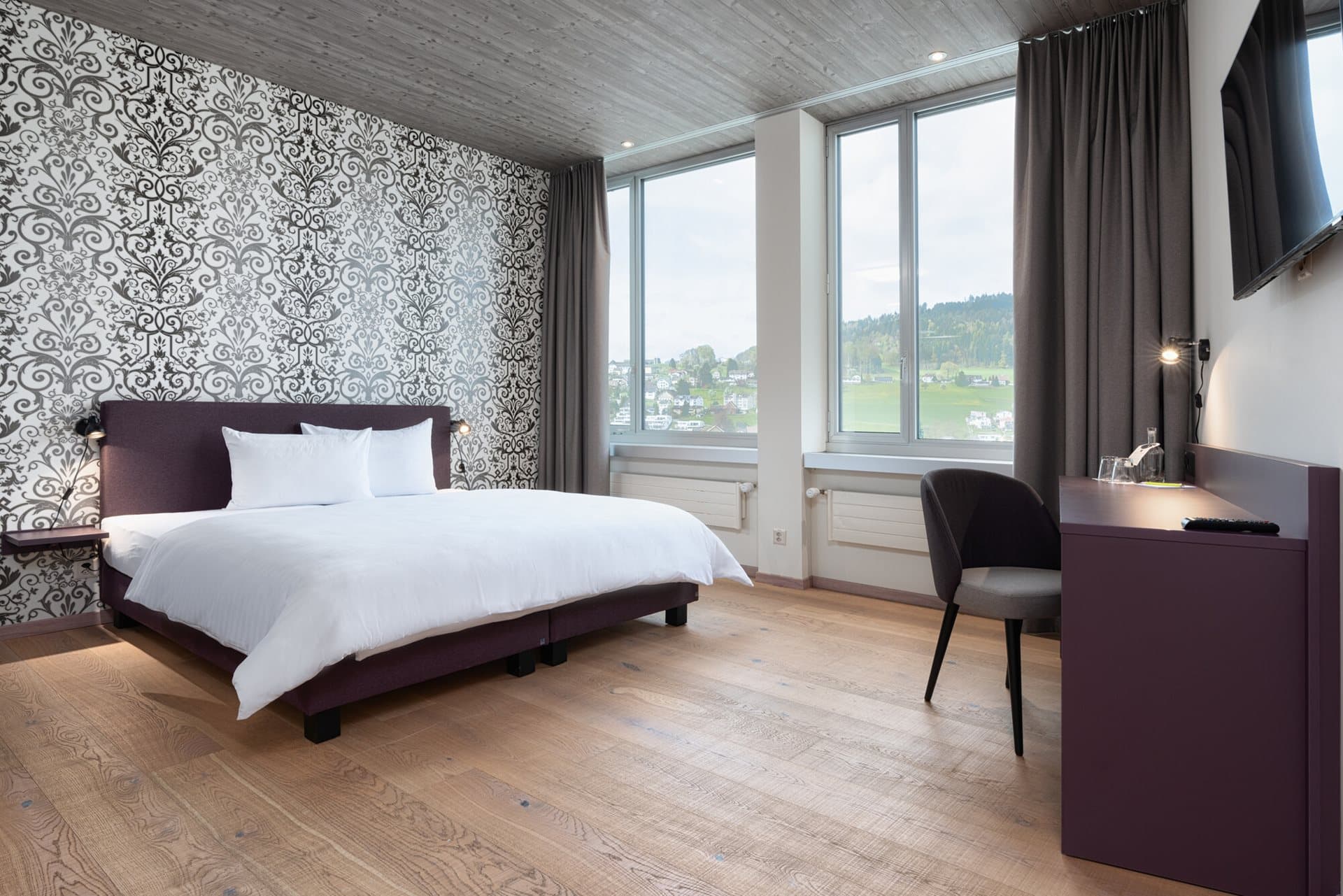 A stylishly furnished hotel room at the Bsmart Hotel Menziken with a modern bed, hardwood floor, designer wallpaper, and a desk with a view of the green landscape.