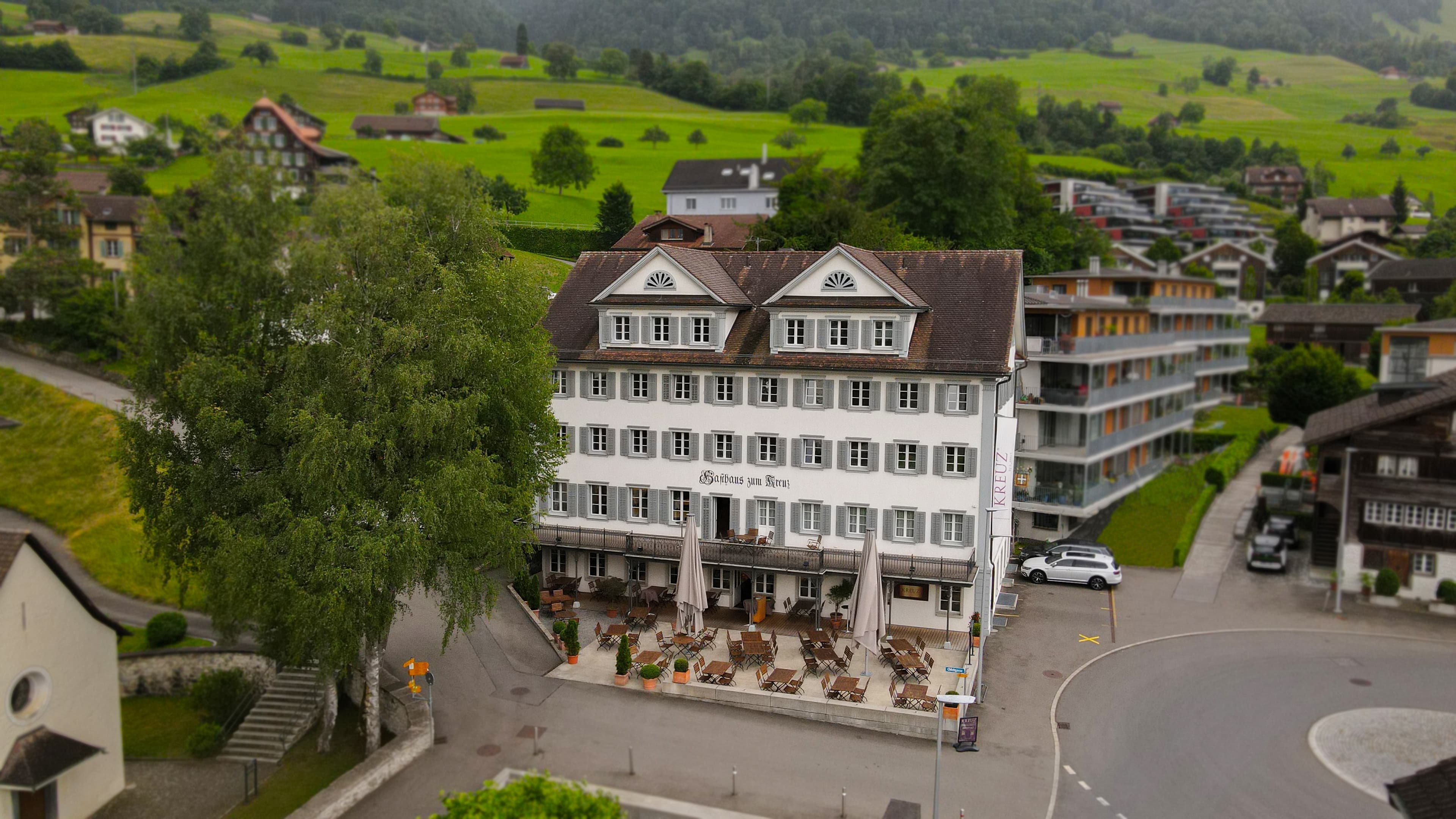 The Inn at the Cross in Sachseln, Switzerland, is a multi-story historic building with a terrace, surrounded by green landscapes and traditional houses.