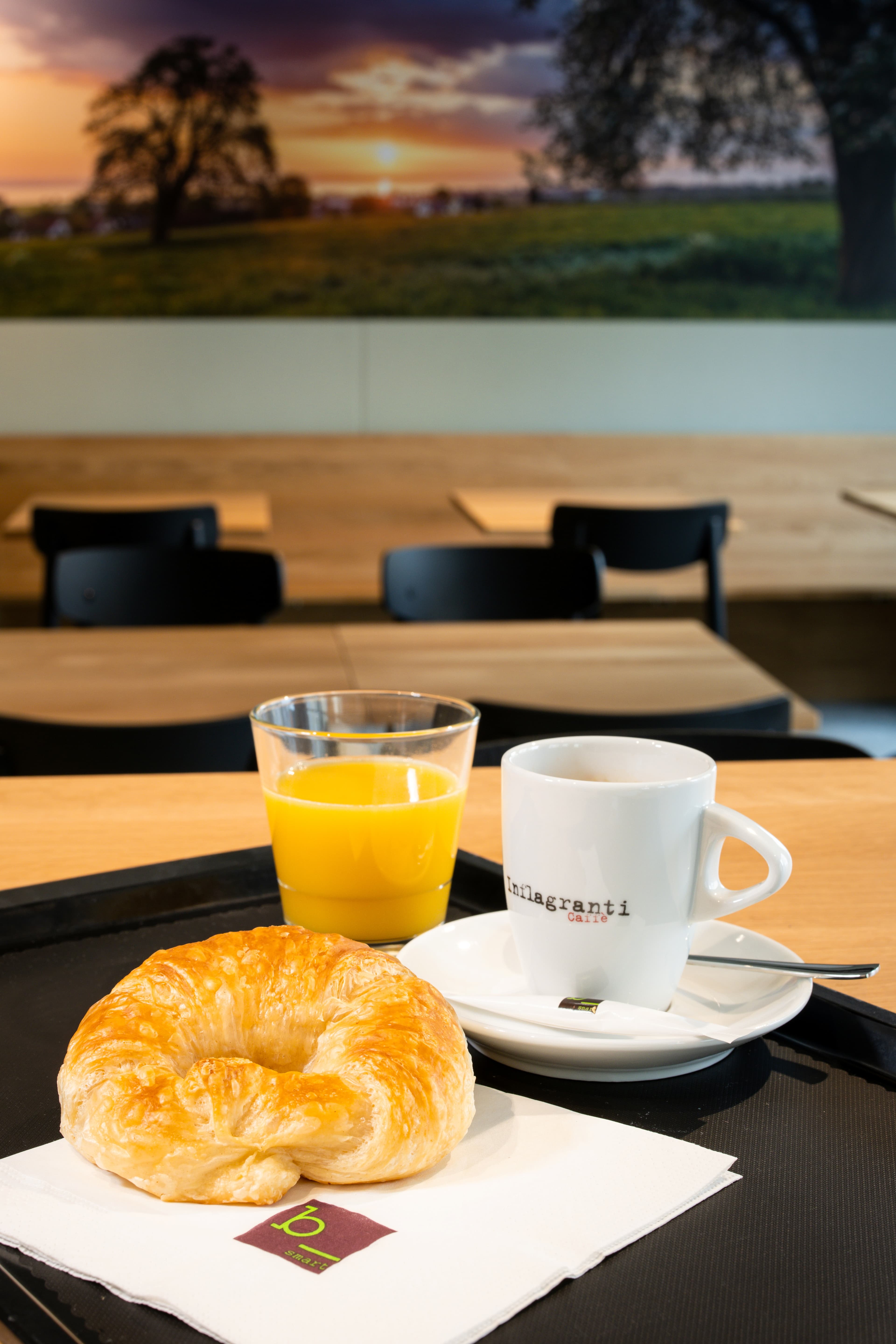 A tray with a croissant, a glass of orange juice, and a cup of coffee at the B_smart Motel Amriswil, with a sunset panorama in the background.
