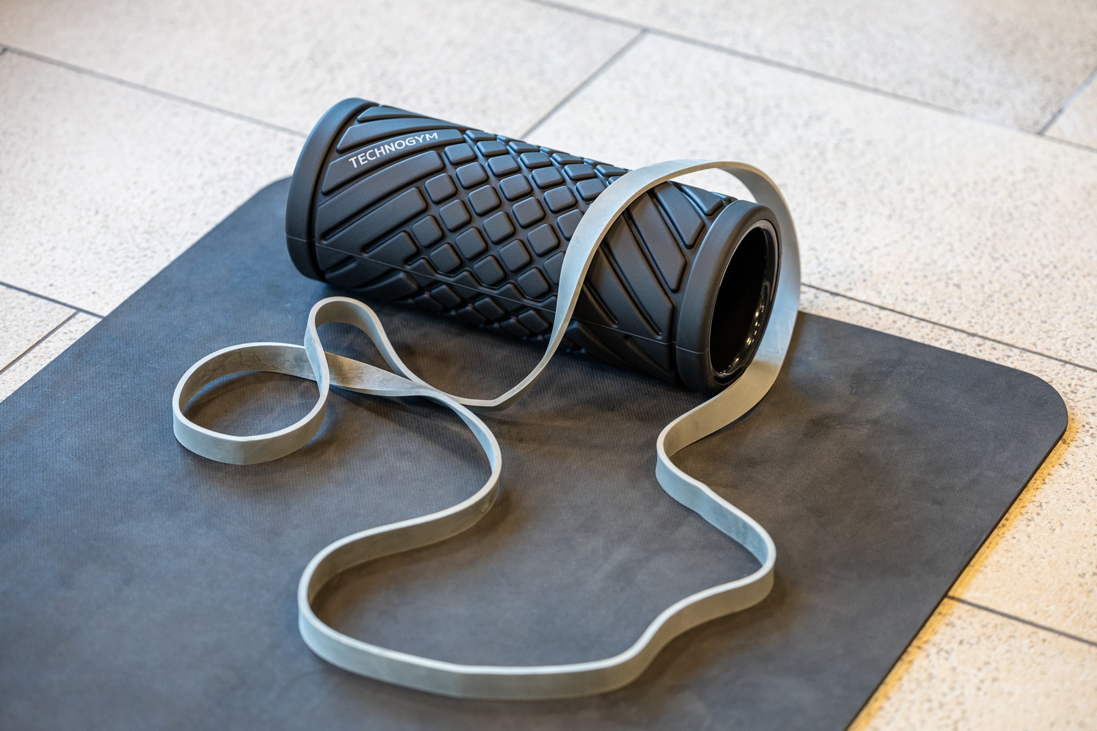A foam roller from Technogym and a resistance band are lying on a mat in a gym.