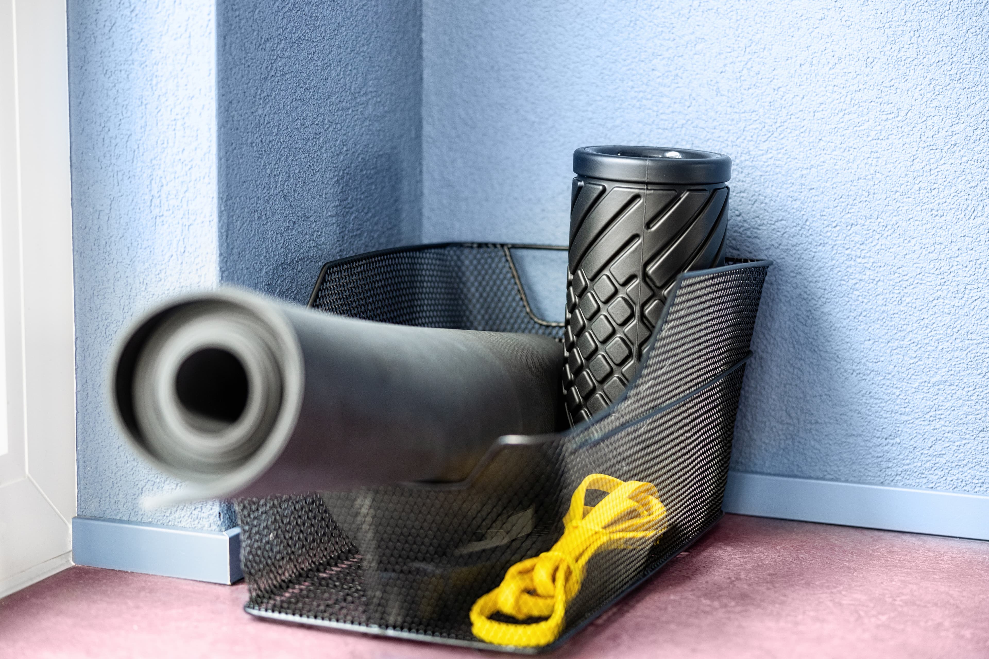 A black exercise mat, a black foam roller, and a yellow jump rope are in a black basket on a pink floor in front of a blue wall.