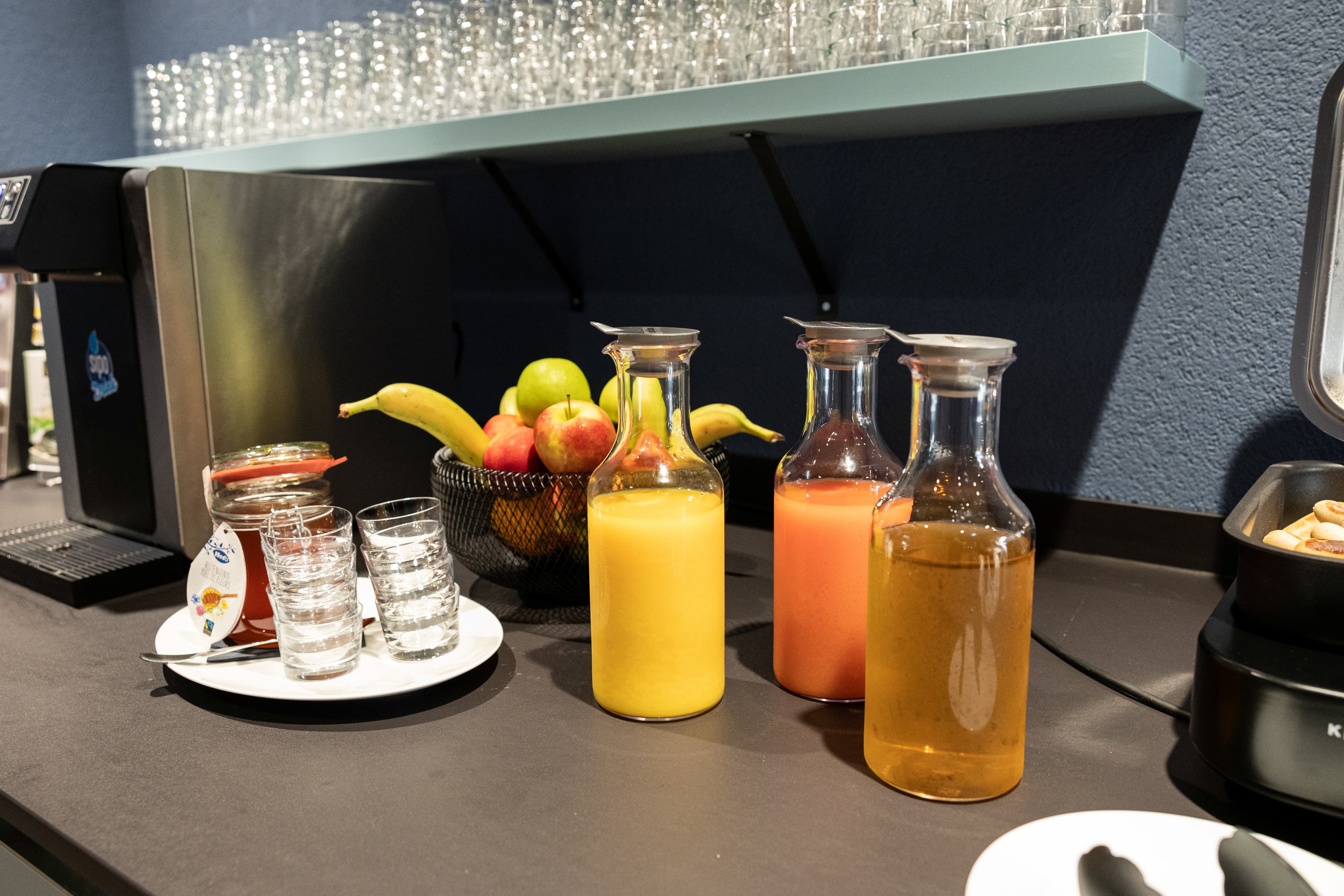 Three glass carafes with different juices and a bowl of fruit are on a breakfast buffet at the B_Smart Hotel Arbon.