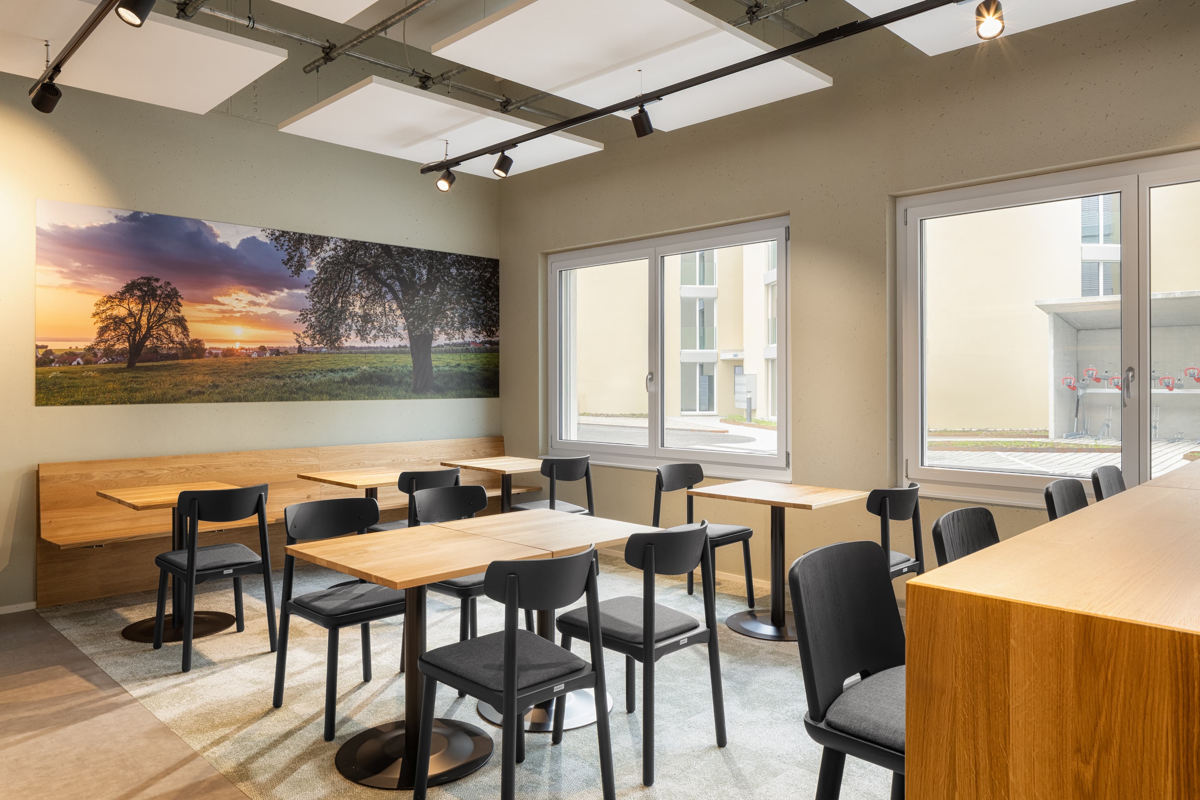 A breakfast area in the B Smart Motel Amriswil with wooden tables and black chairs, decorated with a large landscape picture on the wall.