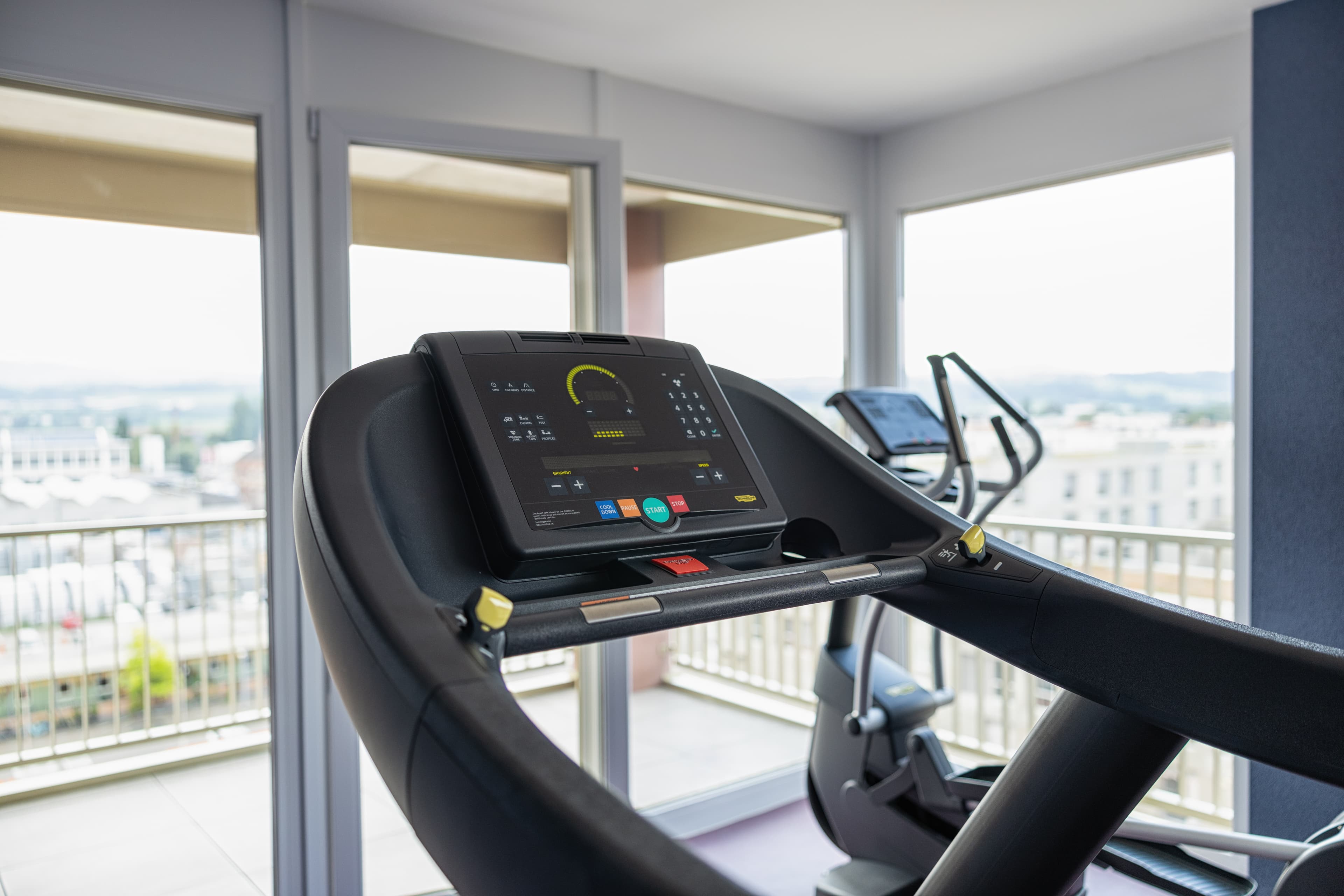 Treadmill in the fitness room of the Bsmart Hotel Arbon with a view of the city through large windows.