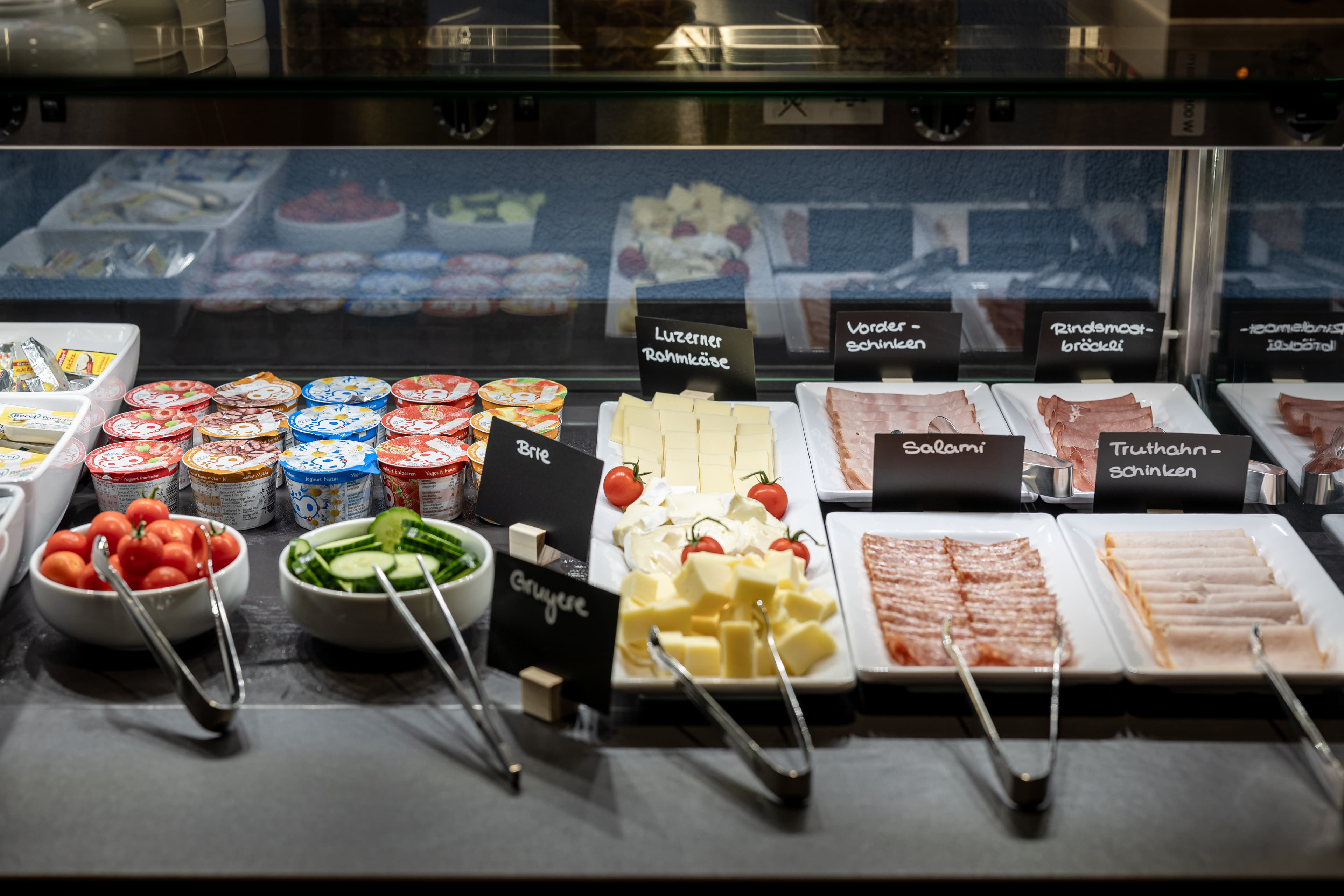 A breakfast buffet at Smart Hotel Arbon with a selection of meats, cheese, yogurt, and fresh vegetables.