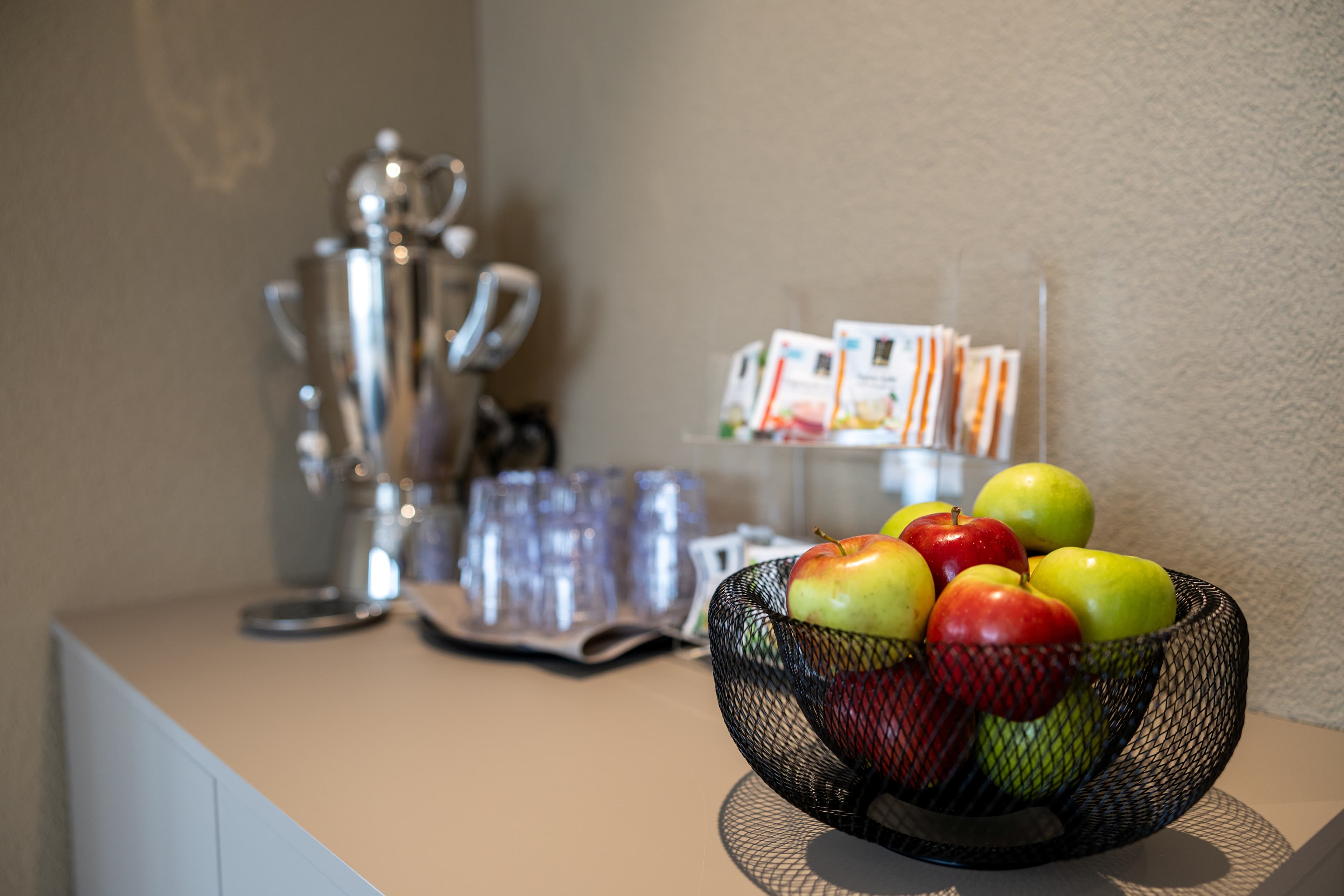 A table in the B smart Hotel Arbon with a bowl full of colorful apples, a samovar, plastic cups, and tea bags.