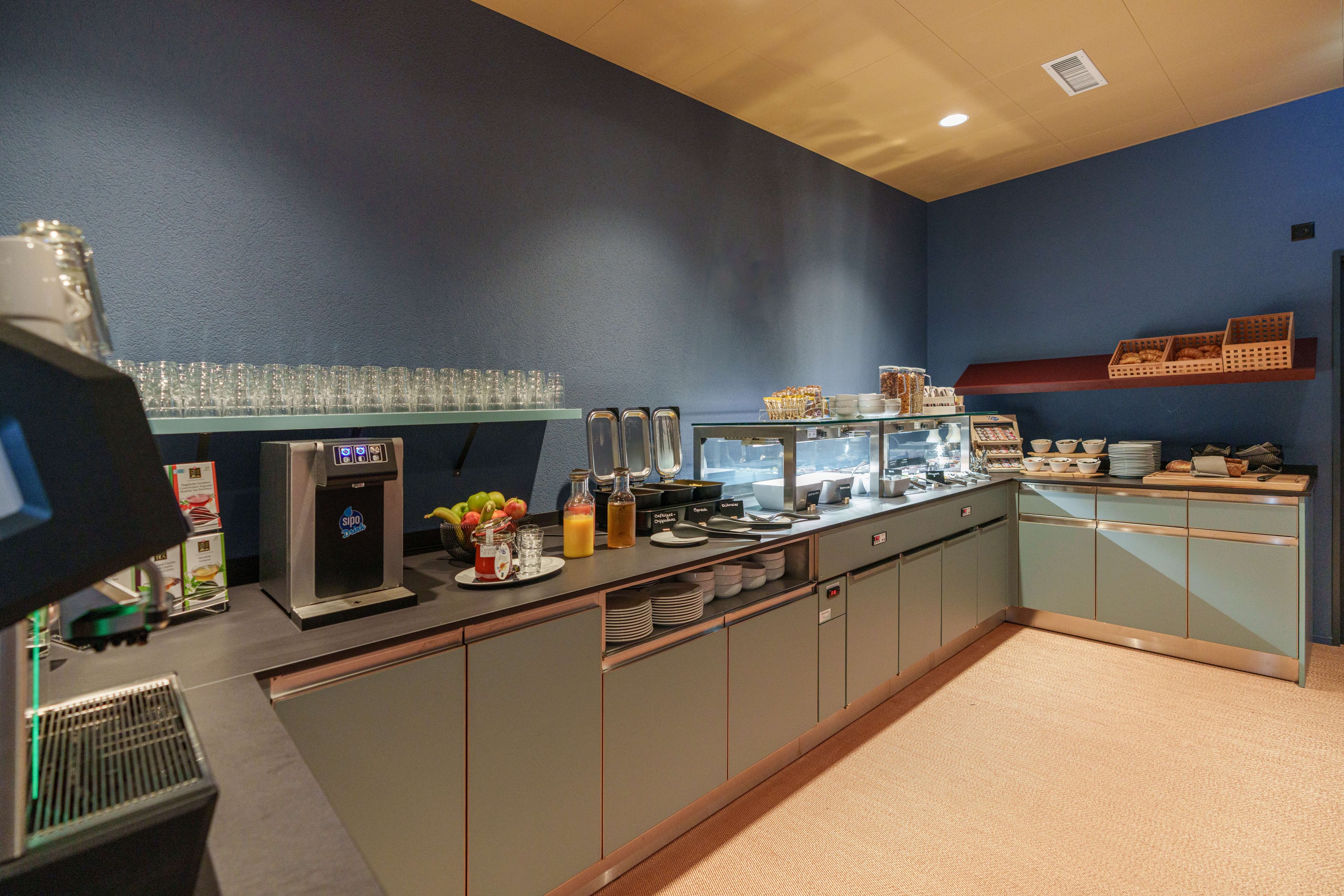 A breakfast buffet at B_Smart Hotel Arbon with rolls, fruit, drinks, and dishes on a modern counter.