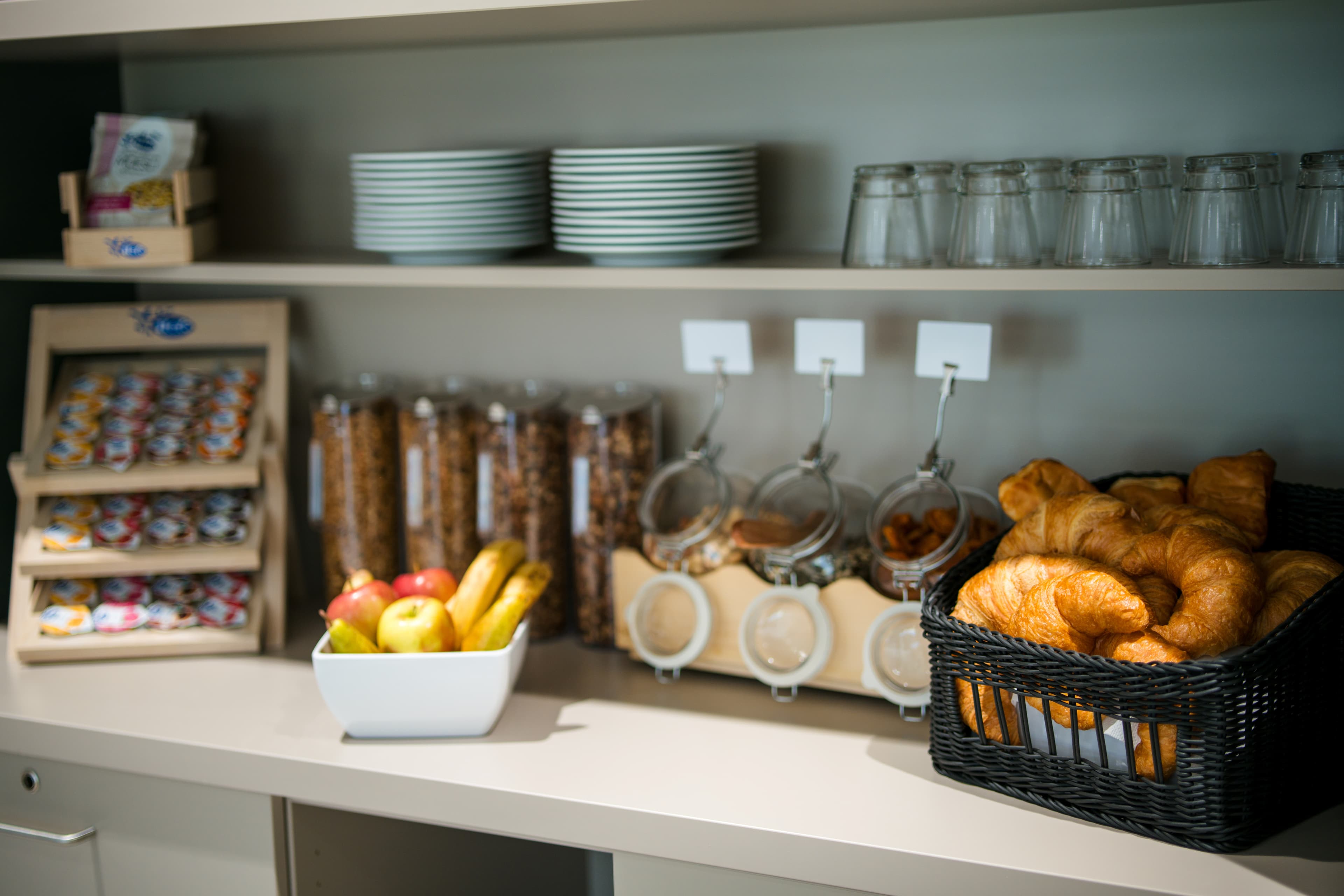 A breakfast buffet at the B_smart Motel Schaan with croissants, fresh fruits, cereal in dispensers and plates, cups, and jams