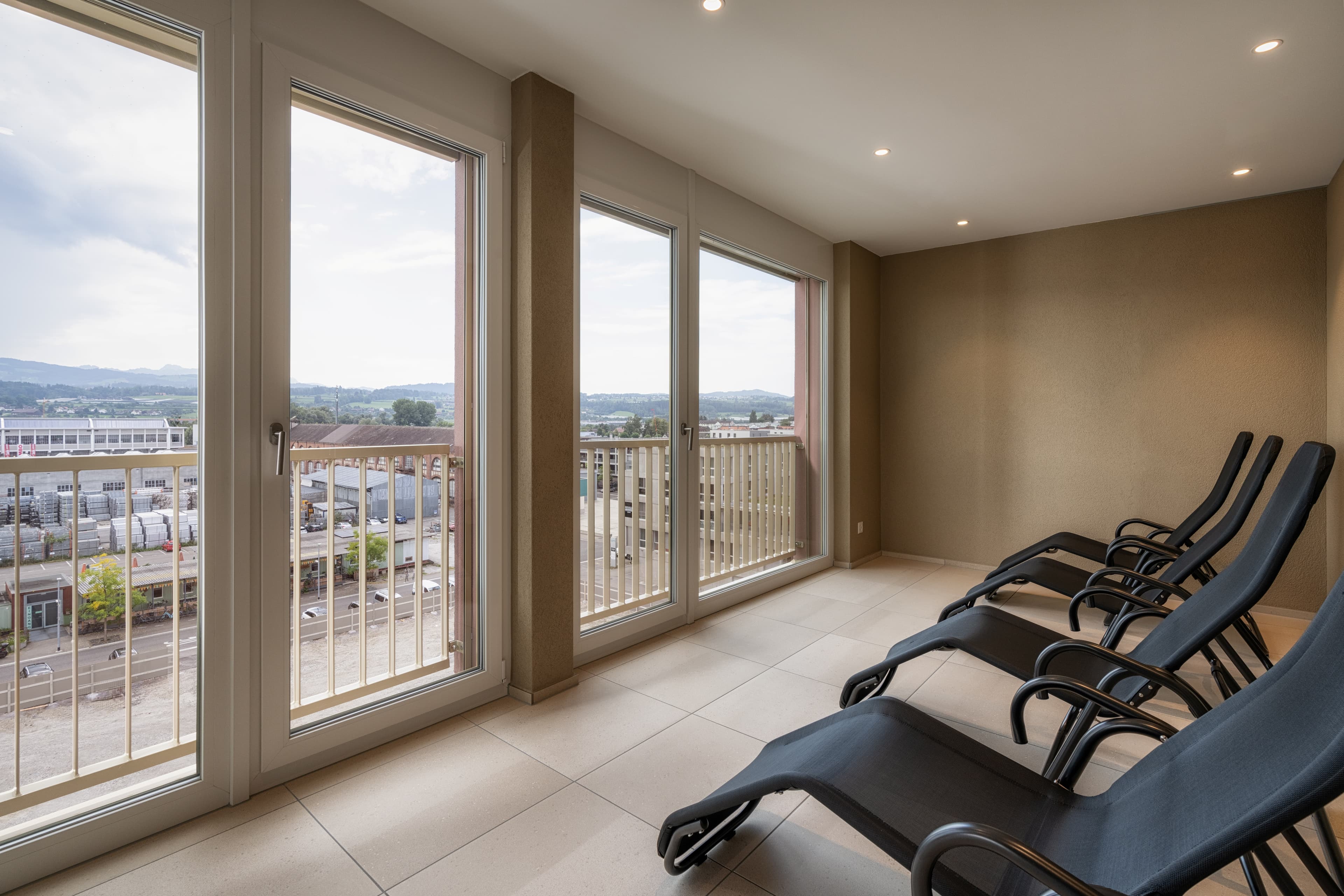 A bright room with lounge chairs at the B_smart Hotel Arbon offers a view of the surrounding city and landscape through large windows.