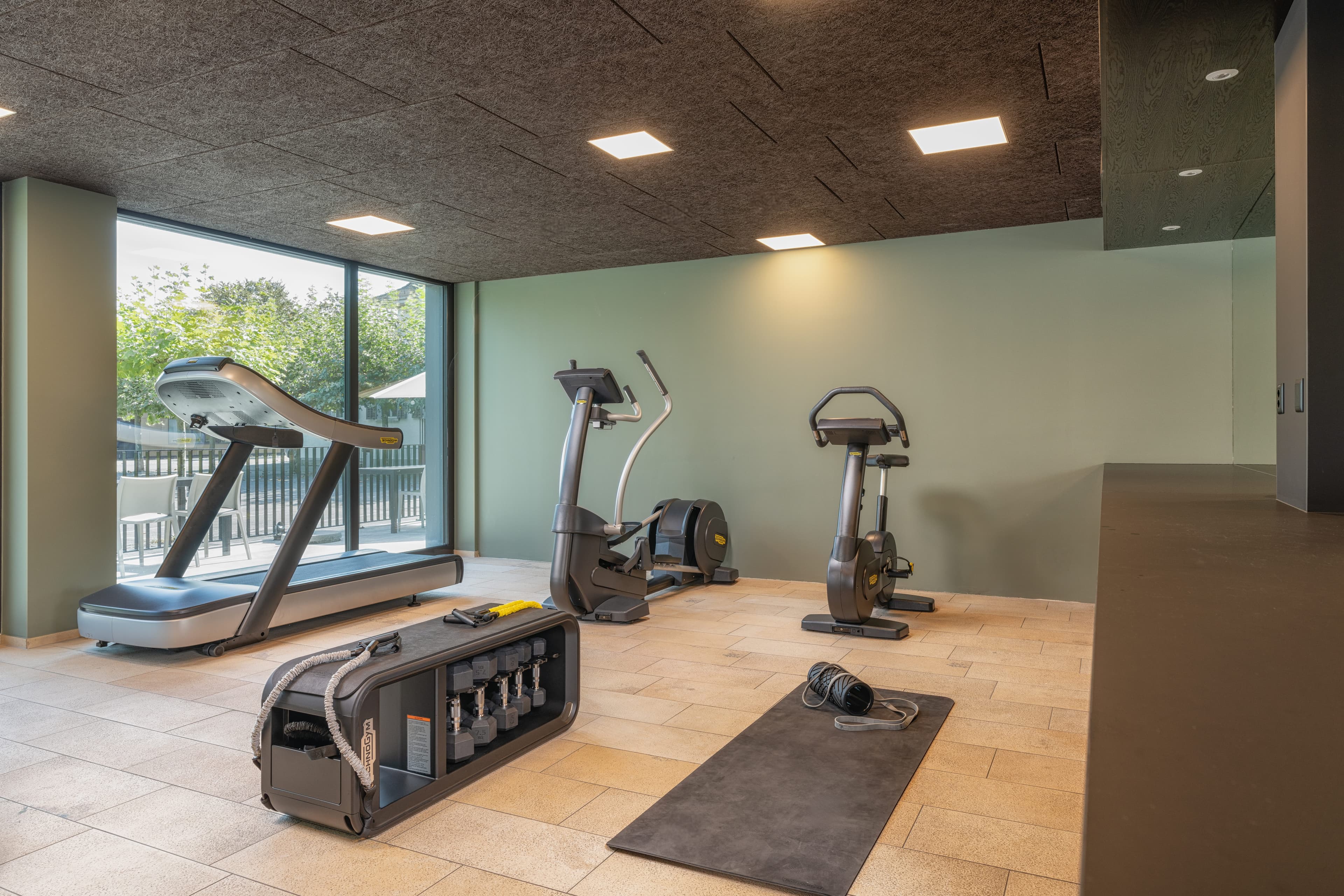 A fitness room in B_smart Hotel Widnau with modern exercise equipment such as treadmill, elliptical trainer, exercise bike, and free weights in front of large windows.
