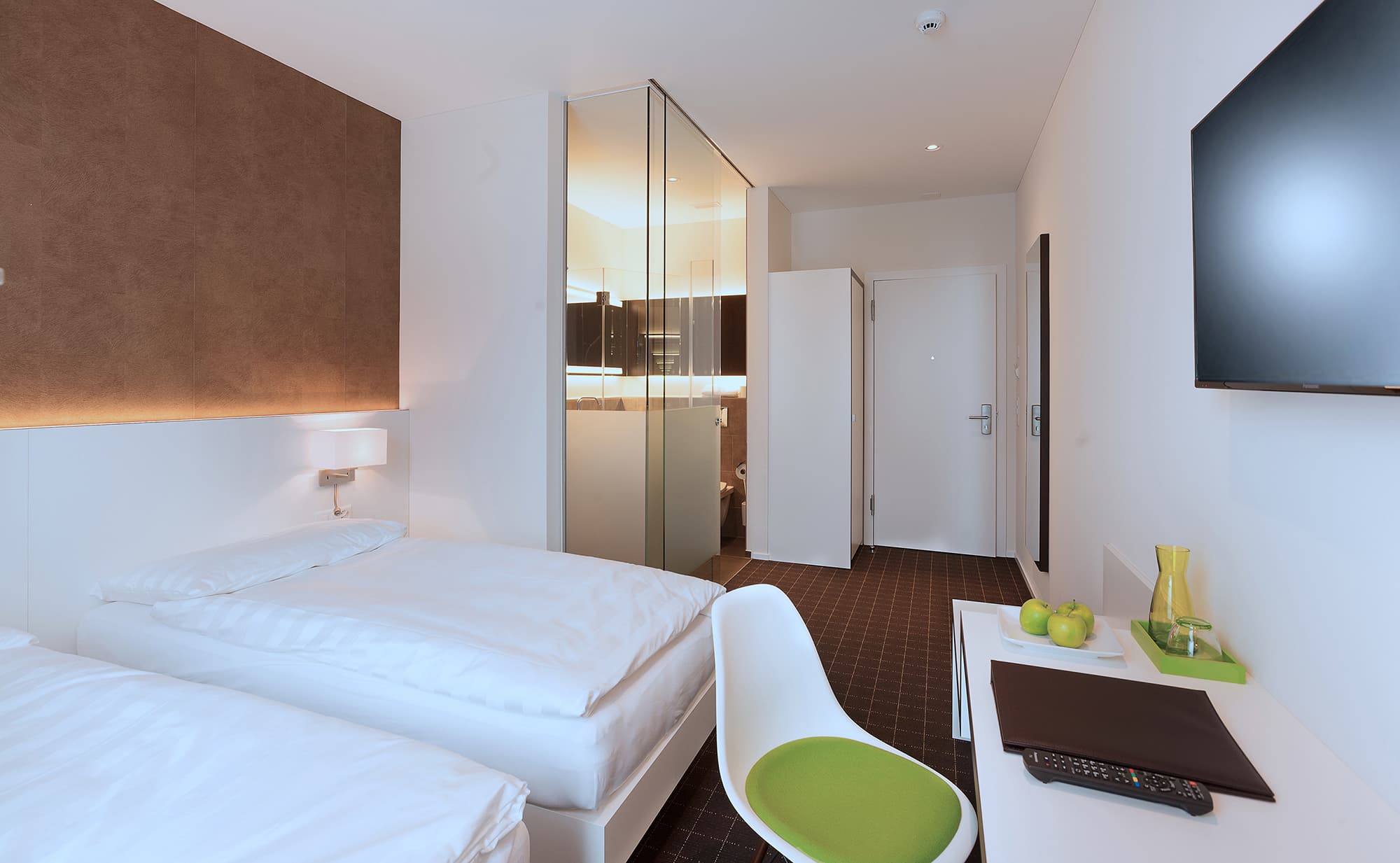 A modern double room in the B Smart Motel Sevelen with two twin beds, a desk with green decor, a TV, and a bathroom with a glass wall.