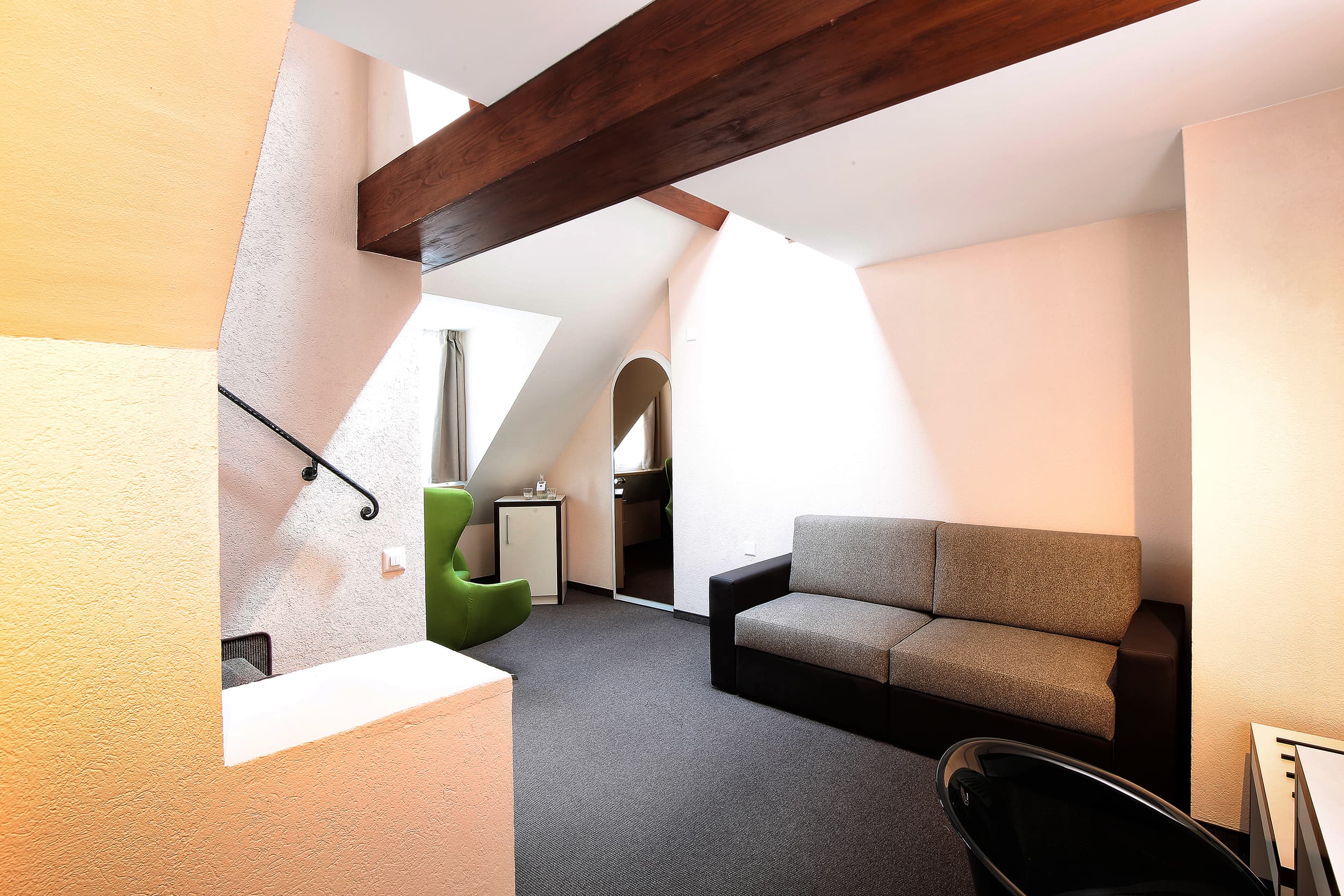 Bright and modern room in a Junior Suite at the B smart Motel Basel with a gray sofa, green chair, and wooden beams on the ceiling.