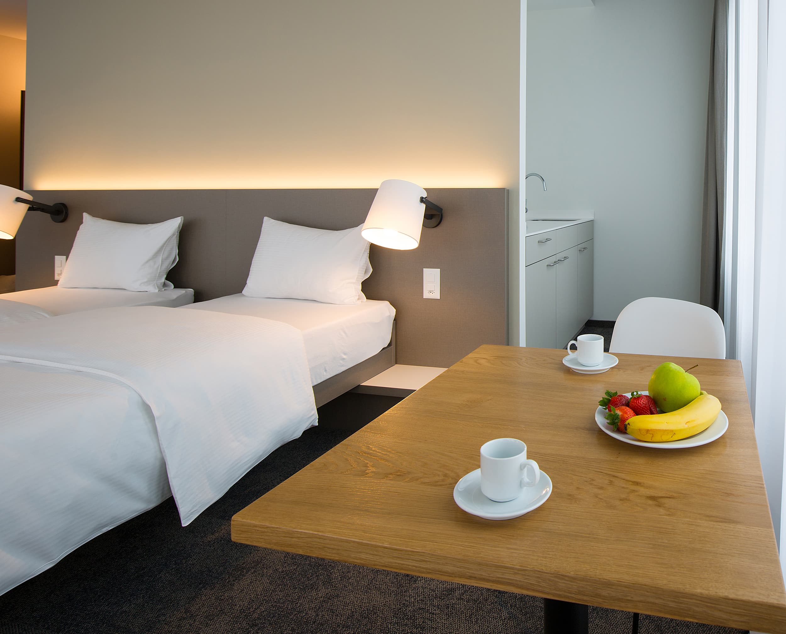 A bright junior suite in the B Smart Motel Landquart with two single beds a table with coffee cups and fruit and a small kitchenette in the background