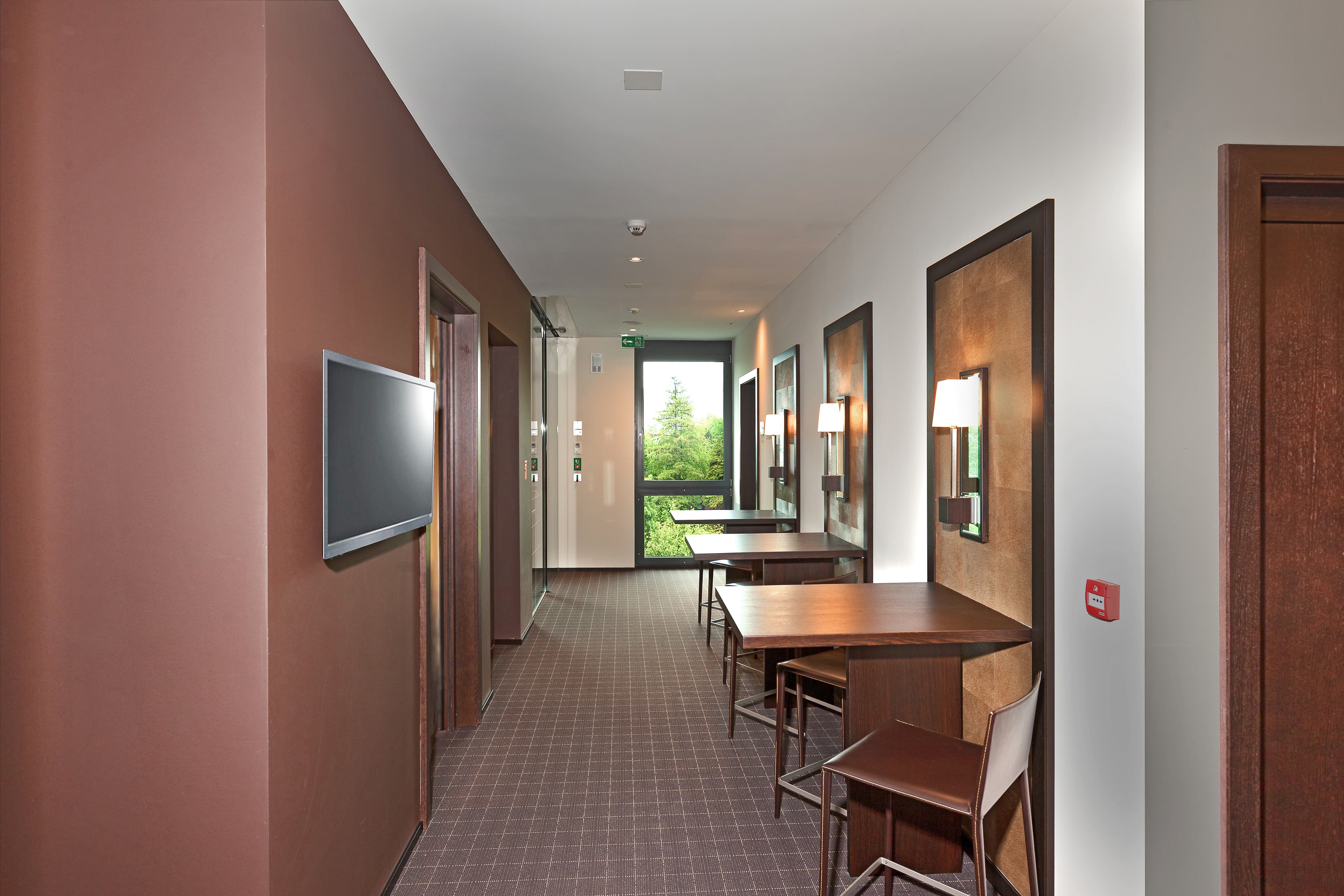 A bright corridor in the b smart motel Sevelen with brown walls, wooden desks and chairs, features decorative wall lamps and a window with a view of greenery.