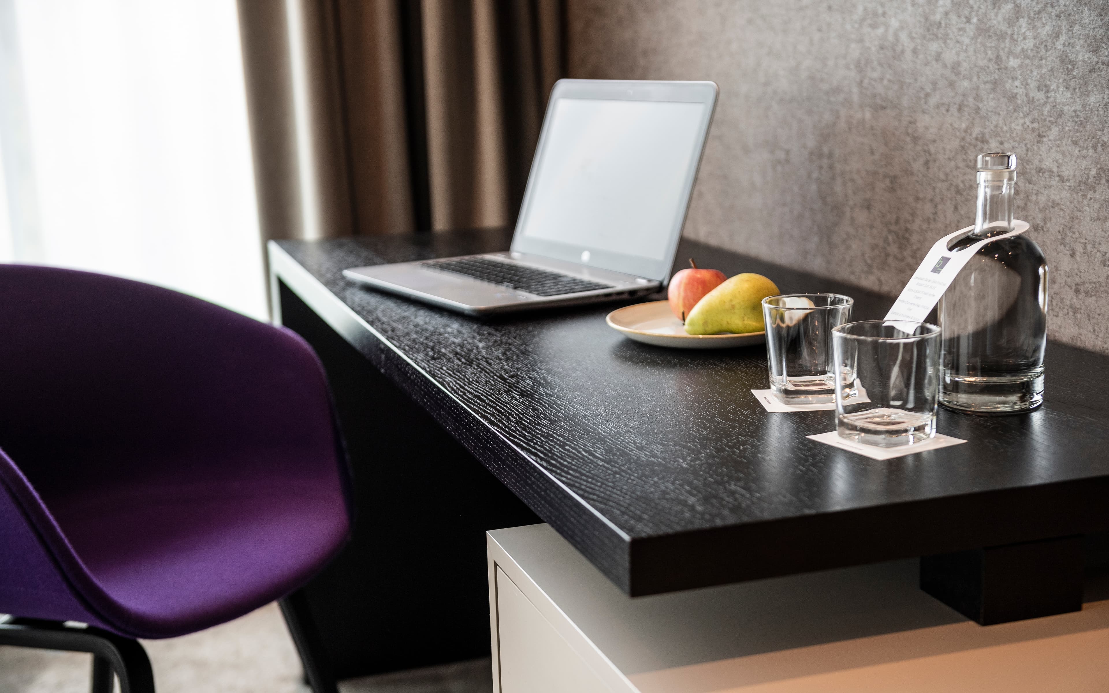 A hotel room at the B_smart Hotel Bendern with a desk on which there is a laptop, a water bottle, two glasses, and a plate with fruit.