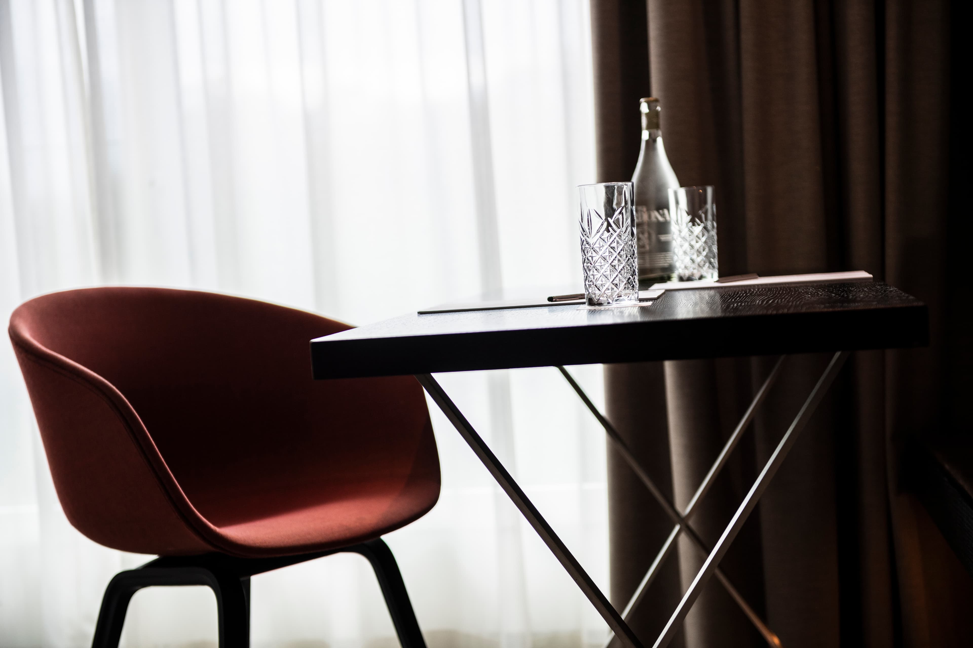 A modern hotel room in the B Smart Hotel Bendern with a dark table, two crystal glasses, a water bottle, and a red chair in front of a large window with curtains