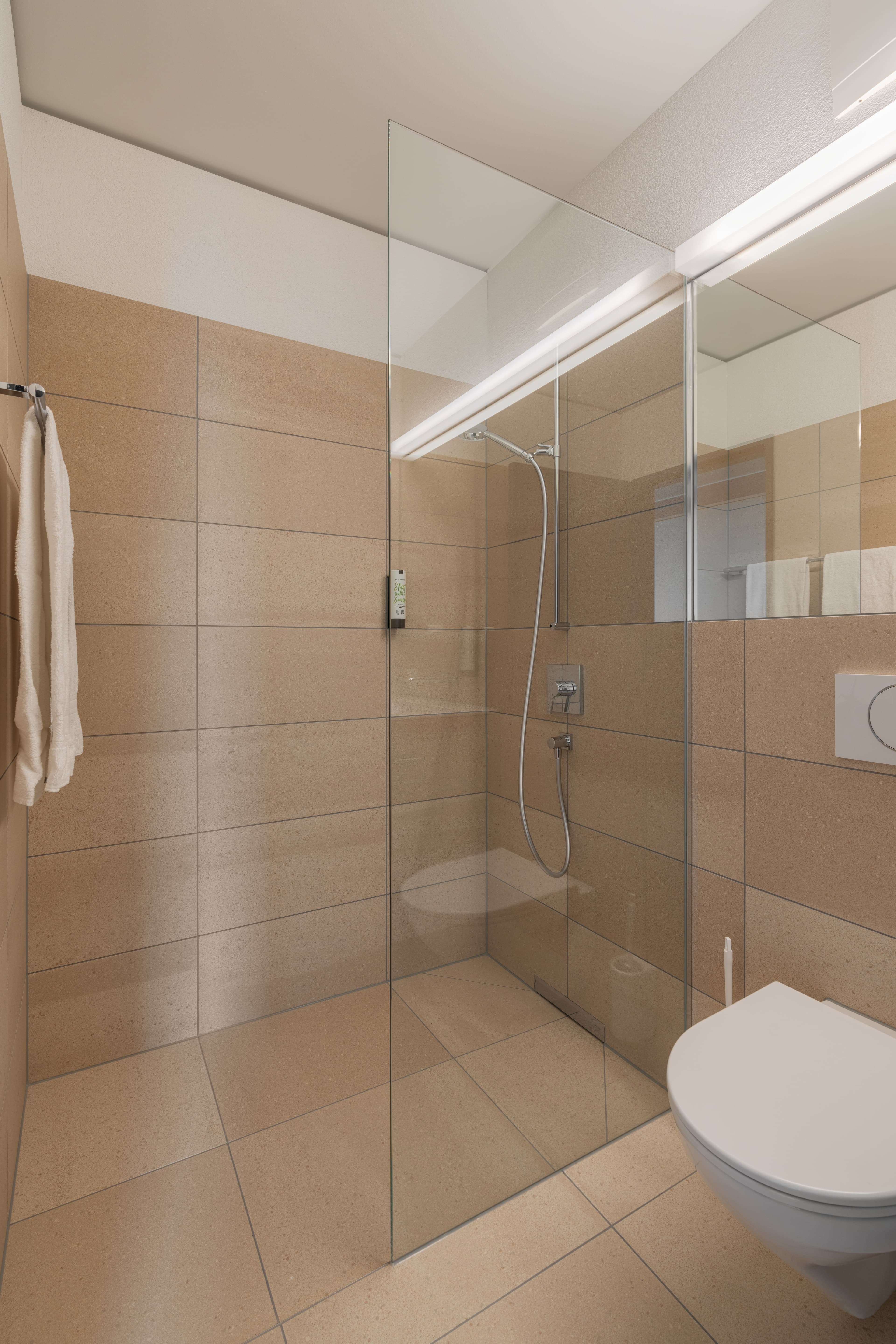Modern bathroom in a Superior Double Room at the B Smart Hotel Arbon with beige tiles, a glass shower, and a wall-mounted toilet.