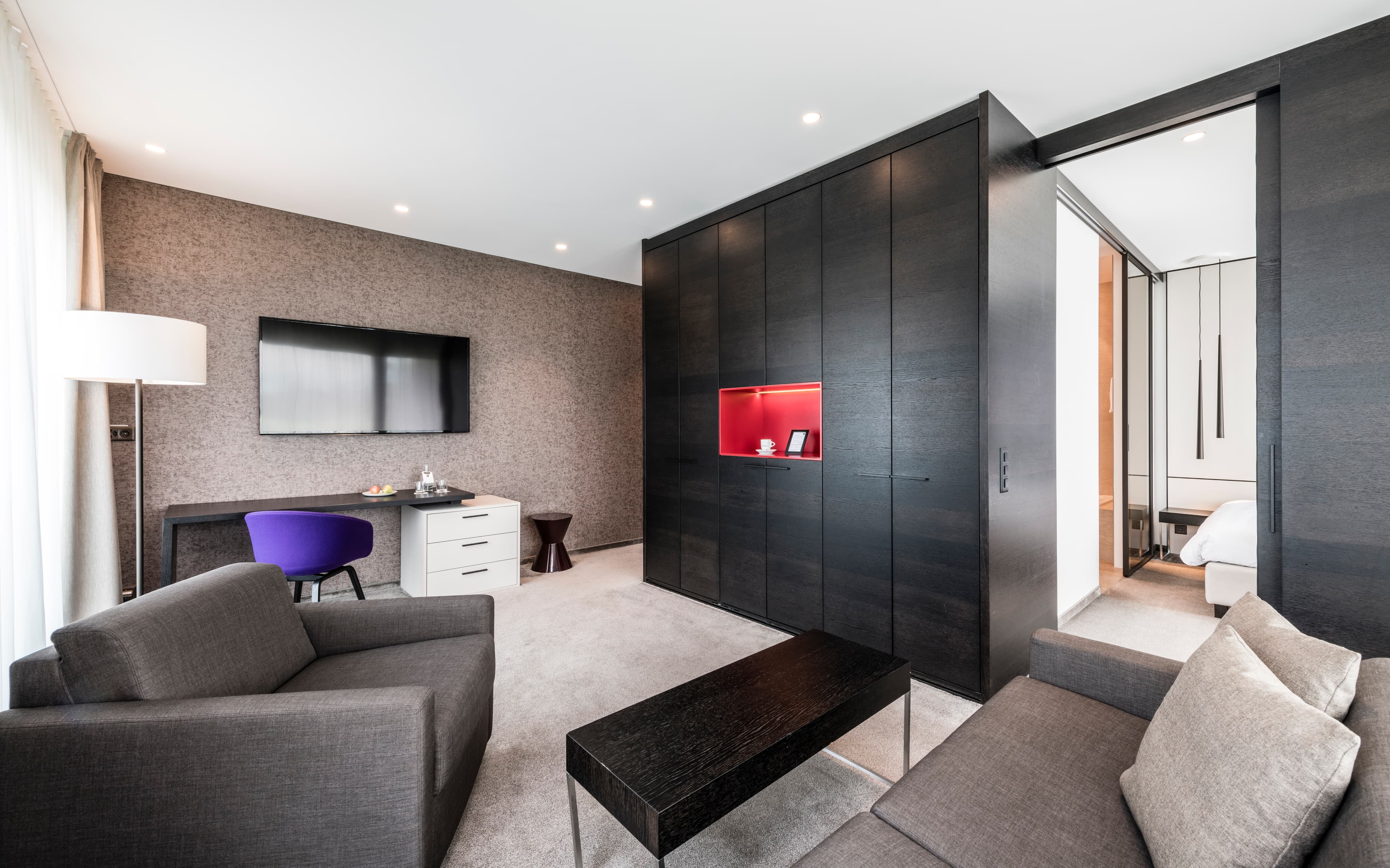 A modern hotel suite with a sofa, armchair, large wardrobe, desk, flat screen TV, and an adjoining bedroom at the b smart hotel in Bendern.