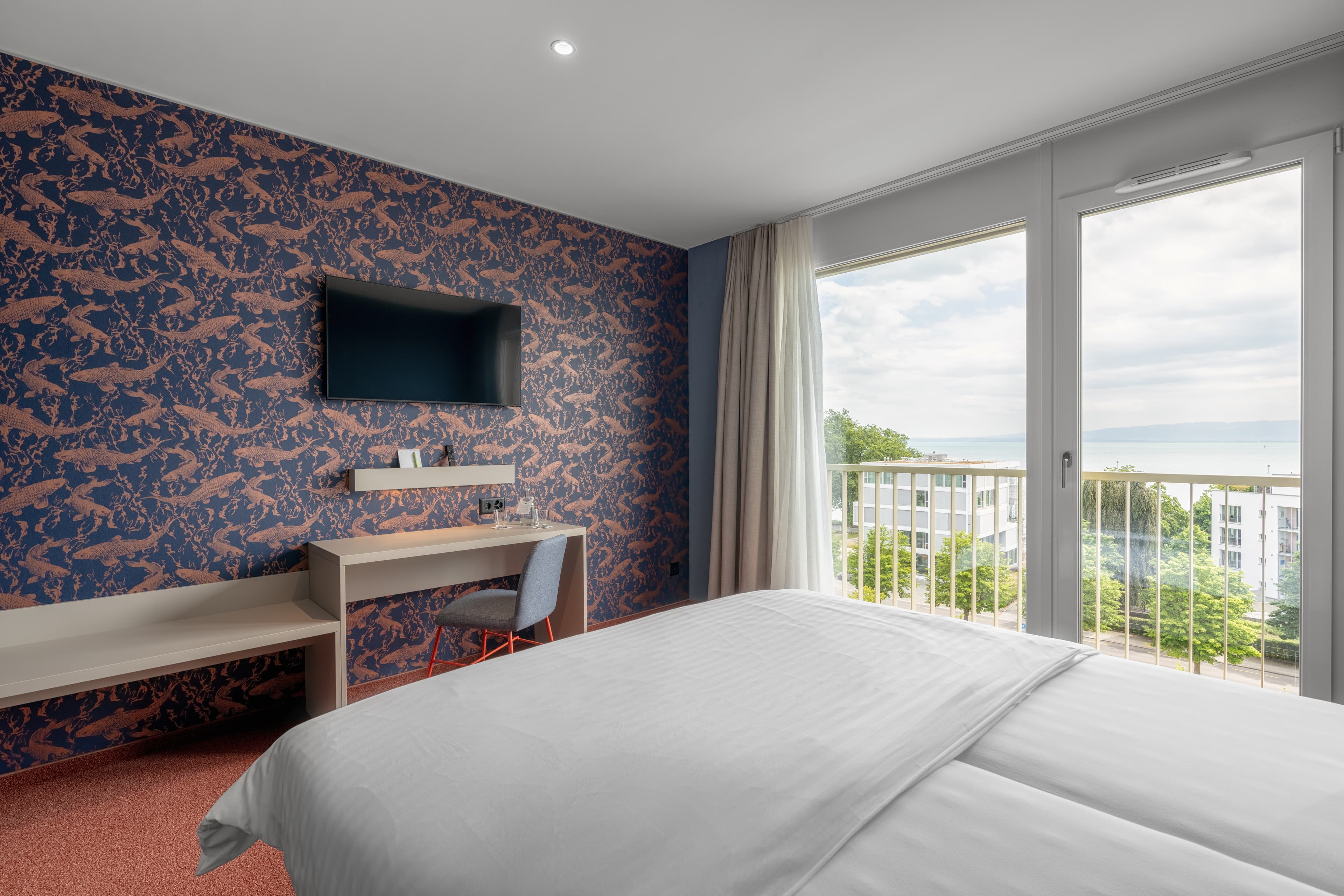 In a bright hotel room with a large window and a view of the lake there is a double bed as well as a desk with a chair and a TV on a patterned wall