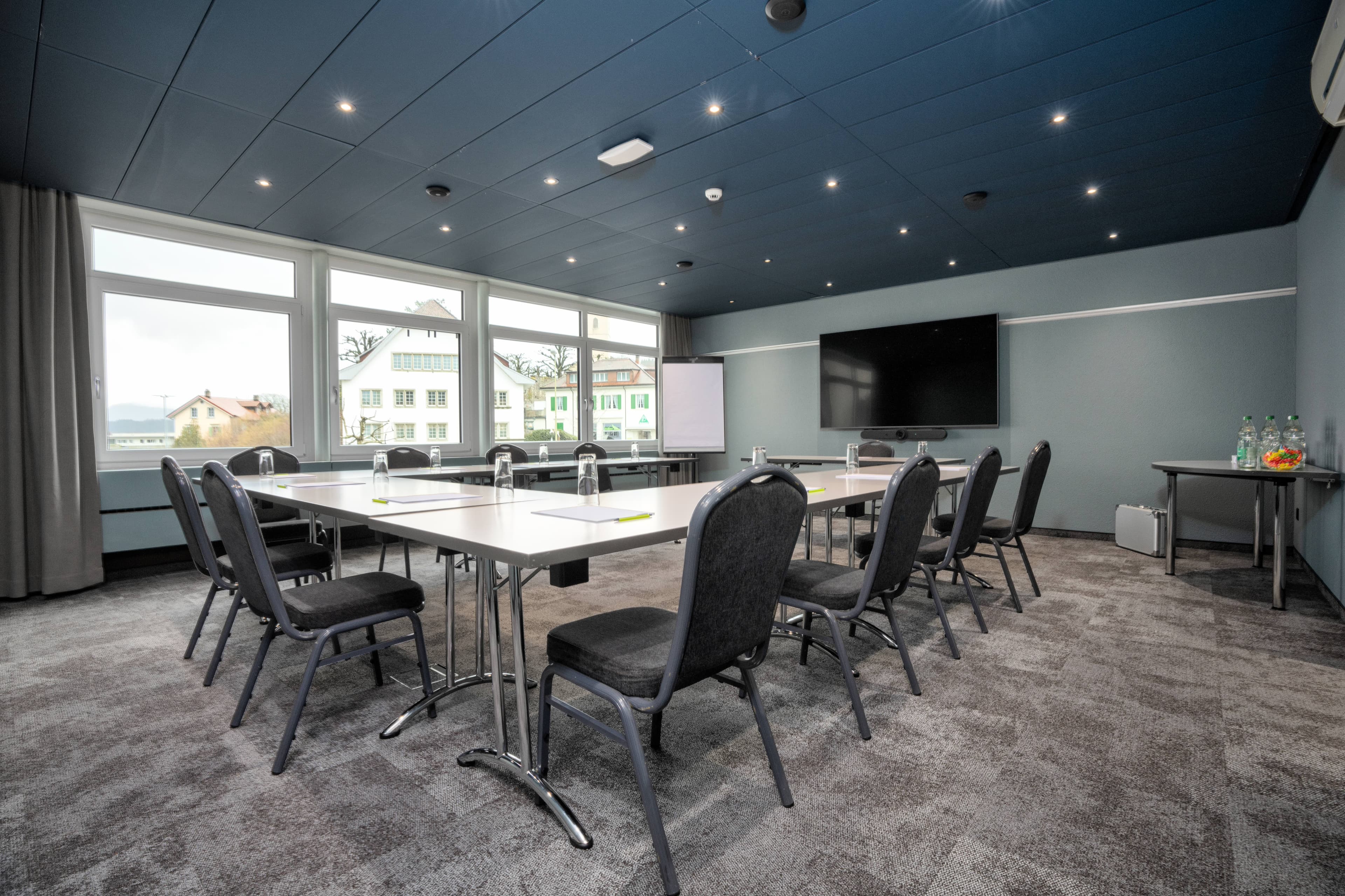 A modern meeting room in the B_smart Hotel Schönenwerd with rectangular tables, chairs, a large screen on the wall, and panoramic windows with a view outside.