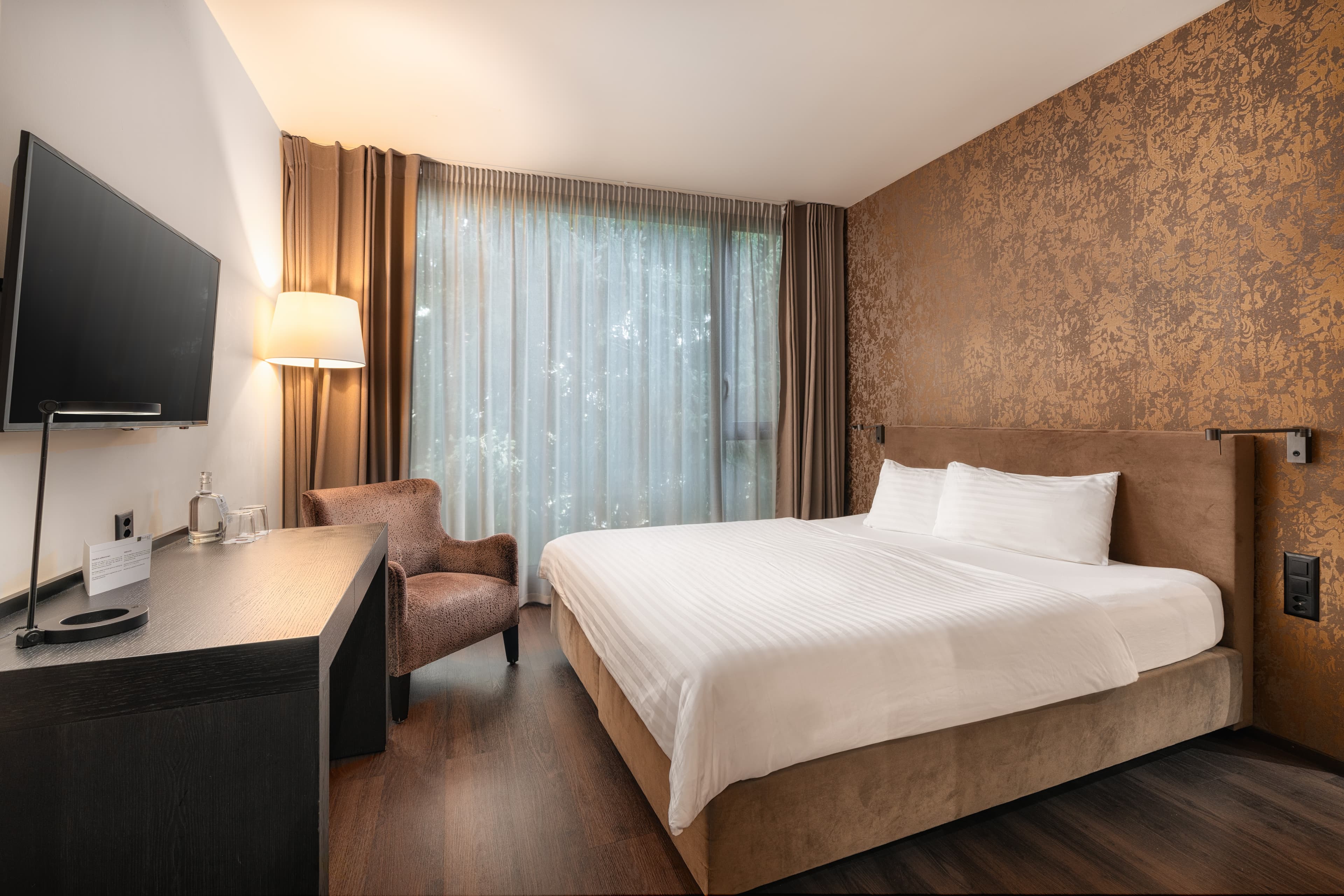 Standard double room at the B_Smart Hotel Widnau with a large bed, desk, armchair, flat screen TV, lamp, and curtains in front of a window.