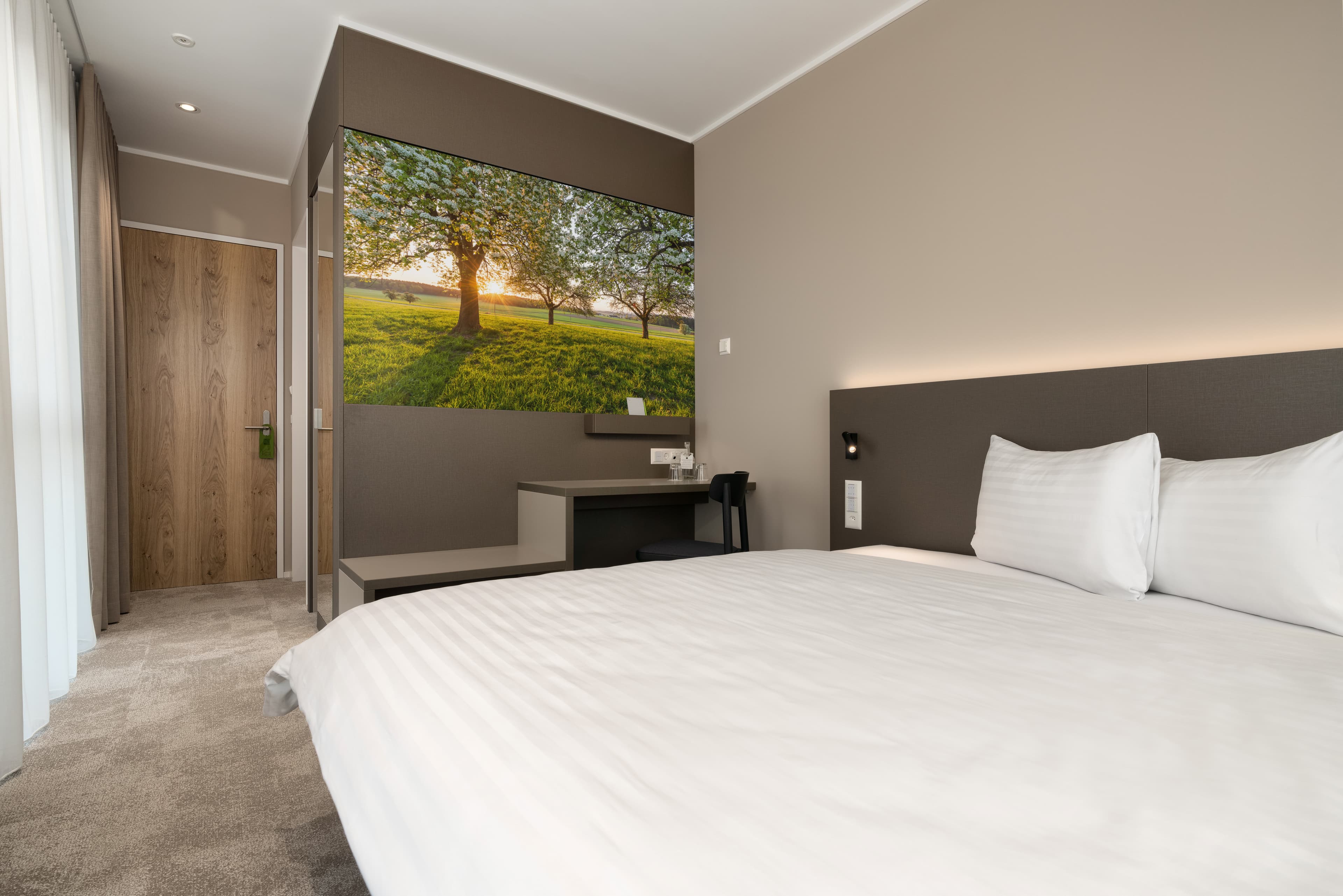 Comfort double room at B Smart Motel Amriswil with a large bed a desk and a wall painting of a tree on a green meadow